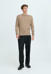 Maglione lavorato a maglia color marrone chiaro con collo rotondo, abbinato a pantaloni neri plissettati e scarpe nere. Design semplice e lineare senza motivi.
