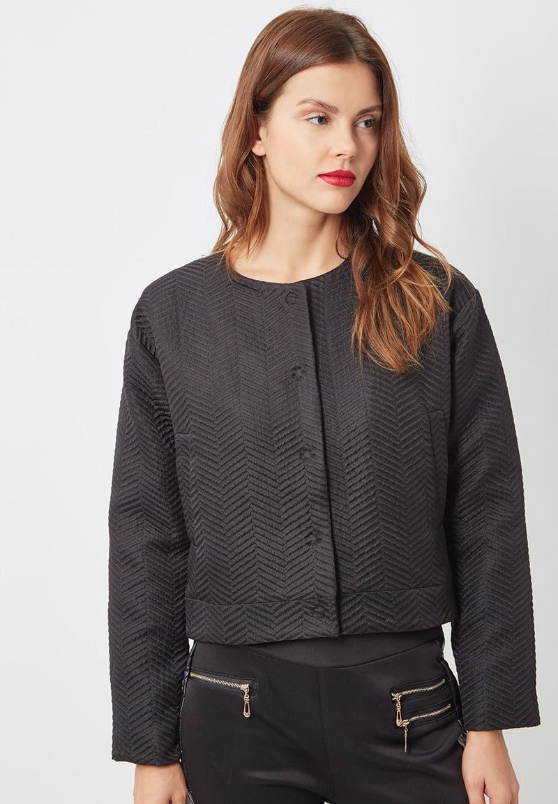 Mujer con cabello ondulado castaño y rojizo, vestida con una chaqueta negra texturizada corta y pantalones negros con bolsillos con cremallera dorada, mirando hacia la derecha sobre un fondo blanco.