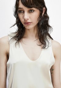Top in satin color crema chiaro con scollatura a V profonda e spalline sottili; texture liscia con una leggera lucentezza.