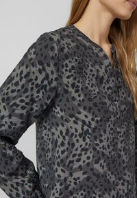 Szara bluzka o luźnym kroju, z czarnym wzorem w kropki, dekoltem w serek i długimi rękawami. Wykonana z lekkiego, teksturowanego materiału.