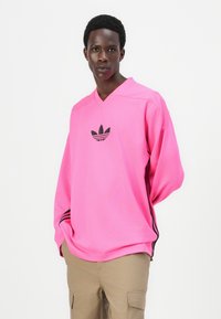 Långärmad V-ringad tröja i klart rosa nätmaterial; har svart Adidas-logotyp på bröstet och svarta ränder längs sidorna.