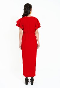 Rochie maxi roșie cu mâneci scurte și vaporoase, talie strânsă și siluetă dreaptă, având un fermoar la spate. Tocuri platform negre.