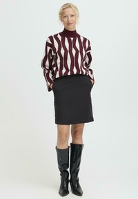Mujer de pie con un suéter de cuello alto de patrón marrón y blanco, falda negra y botas de cuero negras hasta la rodilla, con las manos en los bolsillos.