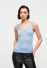 Νεαρή γυναίκα με κοντά σκούρα μαλλιά, φοράει ανοιχτό γαλάζιο τοπ με δαντέλα και μαύρο δερμάτινο παντελόνι, στέκεται με τα χέρια στις τσέπες.