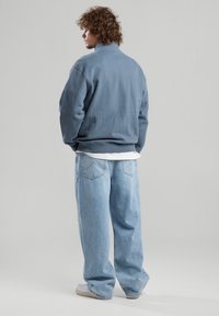 Light blue sweatshirt with a high collar, paired with loose-fitting light blue jeans and white sneakers, showcased from the back.