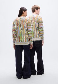 Χρωματιστό πλεκτό πουλόβερ με σπειροειδή σχέδια σε πράσινο, πορτοκαλί και μπλε; λαιμός στρογγυλός; φορεμένο με σκούρο μπλε τζιν παντελόνι με φαρδιά πόδια.