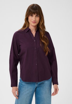 Mulher com cabelo castanho comprido, vestindo uma camisa lilás escura com botões e jeans azuis, em pé contra um fundo claro e simples.
