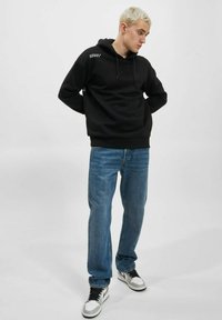 Svart hettegenser med lomme foran, kombinert med blå denimjeans og svarte joggesko med hvite detaljer.