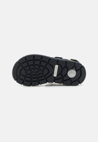 Svart gummisula till sandal med en texturerad, mönstrad yta och greppgropar; har en rund logotypdetalj.