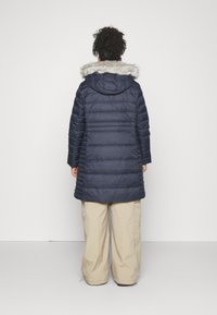 Marineblauer Steppmantel mit Kapuze aus Kunstfell, gesteppter Optik und mittellangem Schnitt. Kombiniert mit hellbeigen, weit geschnittenen Hosen.