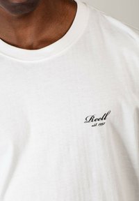 Weißes Baumwoll-T-Shirt mit Rundhalsausschnitt, versehen mit einem kleinen schwarzen gestickten Logo, das auf der linken Brustseite "Reell est. 1997" liest.