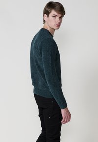 Temno teal pulover z teksturo, dolgimi rokavi in okroglim ovratnikom, v kombinaciji s črnimi hlačami; model obrnjen nekoliko stran od kamere.