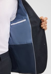 Marineblazer met een gestructureerde buitenkant. Gevoerd met lichtblauwe stof, beschikt over een borstzak en twee binnencompartimenten. Witte stiksels als accenten.