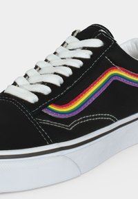 Sort sneakers med hvide snørebånd og sål, med en regnbue-stribe på siden, isoleret på en hvid baggrund.