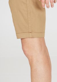 Tan katoen shorts met een rechte snit, omgeslagen zoom en een gladde textuur, zijwaarts getoond op een neutrale achtergrond.