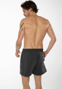Costume da bagno grigio scuro da uomo con vita elasticizzata, tasche laterali e un sottile motivo testurizzato. Tatuaggi visibili sul braccio.