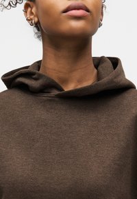 Κοντινό πλάνο του κάτω μέρους του προσώπου και του λαιμού ενός ατόμου που φοράει καφέ φούτερ με κουκούλα, με πολλά ασημένια σκουλαρίκια στο αριστερό αυτί.