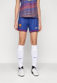 Un short de football bleu avec des bordures rouges et un logo de Barcelone, associé à des chaussettes blanches avec des marques bleues de Nike et des crampons.