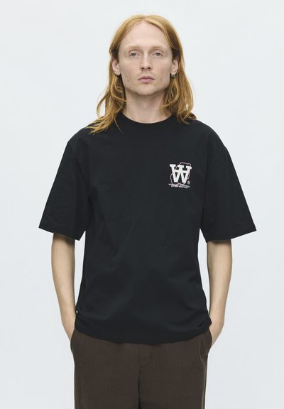Jeune adulte aux longs cheveux roux portant un T-shirt noir oversize avec un petit logo blanc sur la poitrine, debout les mains dans les poches.