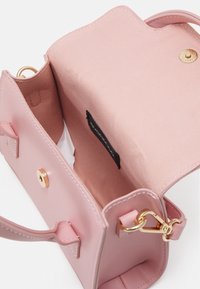 Pieni vaaleanpunainen käsilaukku, jossa on auki oleva läppä, kultainen magneettiklipsi, kultainen rengas ja klipsi, näkyvä kangasvuori ja brändilogo sisällä.