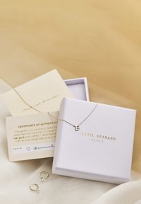 Gouden ketting met een "B" hanger aan een schakel, gepresenteerd in een witte doos. Inclusief een authentiteitscertificaat en gouden creolen met parels.