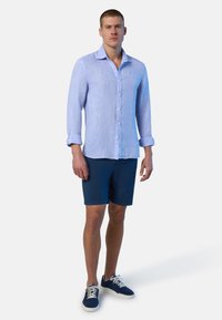 Camicia a maniche lunghe in lino blu chiaro con colletto button-down, polsini arrotolati, abbinata a pantaloni shorts in blu navy e sneakers blu. Design pulito.
