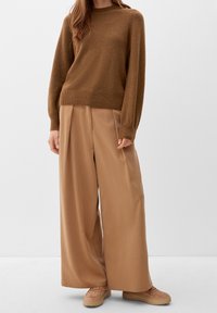 Femme portant un pull en tricot marron, un pantalon large beige et des baskets beiges, se tenant contre un fond clair uni.