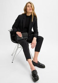 Vrouw met lang blond haar zittend op een zwarte stoel, gekleed in een zwart pak en loafers, met een zwarte handtas met een zilveren ketting en het "HUGO" logo.