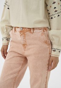 Bēša ar garām piedurknēm krekls ar izšūtām sejām piedurknēs, valkāts kopā ar augsta vidukļa izbalējušām rozā džinsa biksēm stāvošai personai.