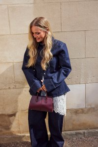 Giacca in denim con maniche ampie di colore blu scuro, indossata sopra un top di pizzo bianco. Tiene in mano una piccola borsa bordeaux con due manici.