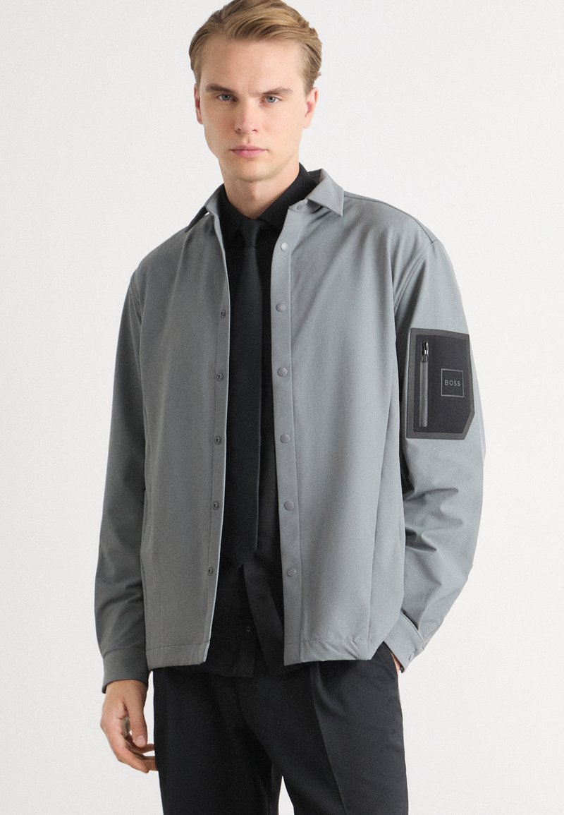 Veste légère grise avec une fermeture à boutons-pression, dotée d'une poche à patch sur le bras gauche ; associée à une chemise noire et une cravate.