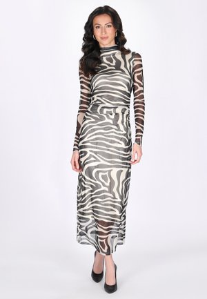 Kobieta w długiej sukience o długości do kostek z nadrukiem w paski zebry, z przezroczystymi rękawami i czarnymi szpilkami, stojąca na białym tle.