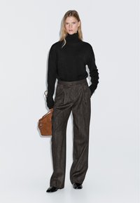 Zwarte coltrui, grijs gestreepte wijde broek en bruine leren handtas. Model staat met handen in de zakken, zwarte schoenen.