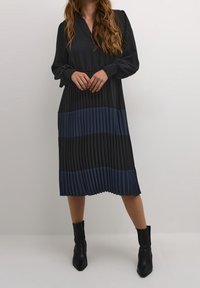 Svart och marinblå plisserad klänning med långa ärmar, krage och smockade detaljer vid handlederna. Ankelhöga svarta stövlar kompletterar outfiten.