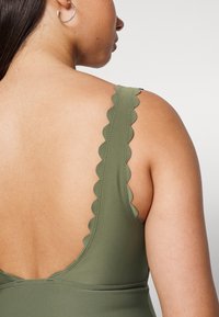 Fato de banho verde oliva com alças em forma de onda, apresentando uma parte de trás baixa e textura suave. Detalhes de costura sutis realçam o design.