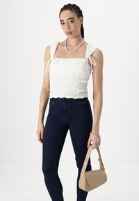 White smocked top with square neckline and tie straps, paired with dark blue skinny jeans. Model holds a light beige shoulder bag.