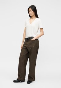 Junge Frau steht mit einer Hand in der Tasche, trägt ein weißes V-Ausschnitt-T-Shirt, braune Hosen mit weitem Bein, schwarzen Gürtel und schwarze Schuhe.