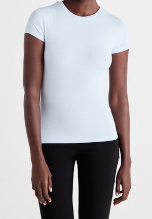 Person iført en tætsiddende lyseblå kortærmet t-shirt og sorte bukser, står mod en ensfarvet baggrund.