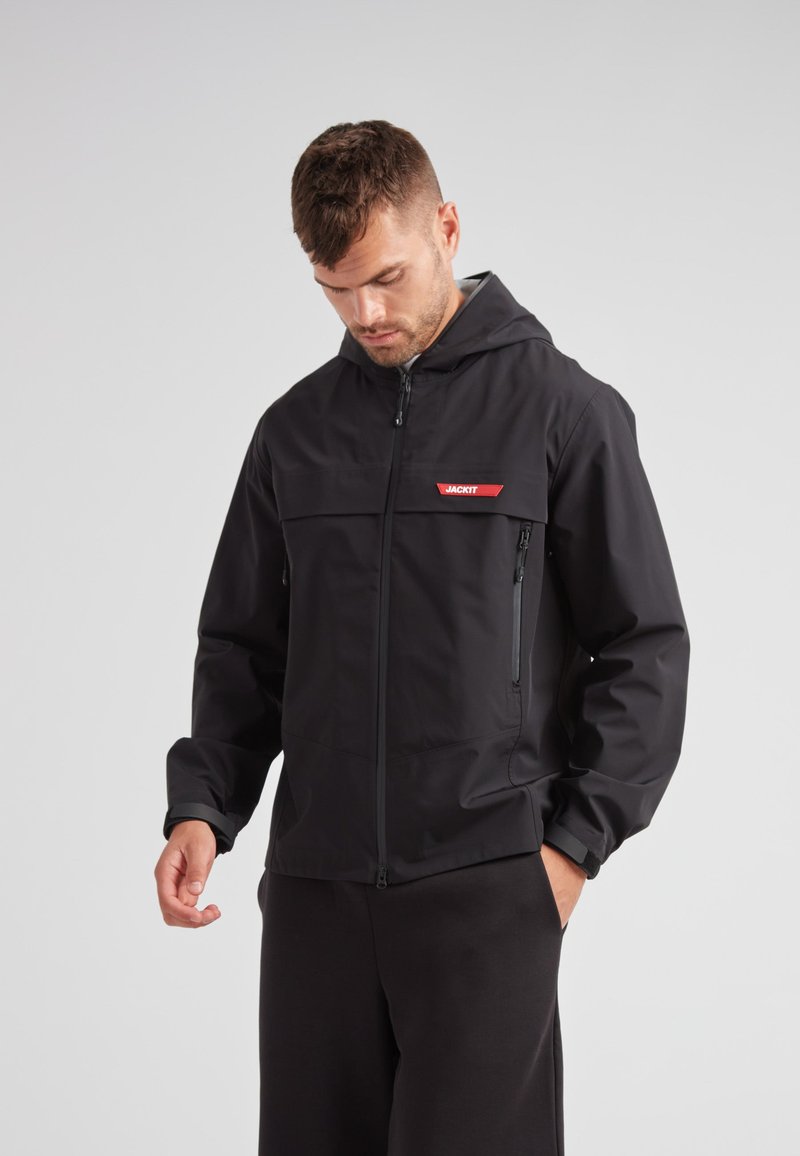 Giacca nera con cappuccio realizzata in un materiale liscio e leggero. Presenta una zip frontale, tasche laterali e un accento con logo rosso sul petto.