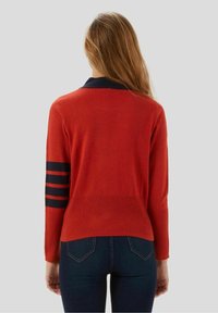 Red sweater with a navy collar and three navy stripes on each sleeve, featuring a textured knit and fitted design. Worn over dark jeans.