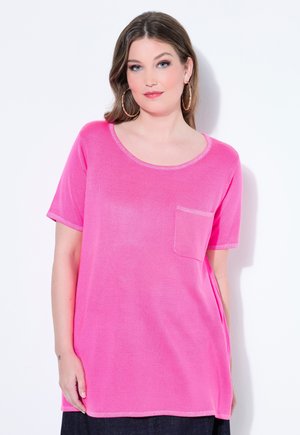 Pitkähiuksinen nainen, jolla on suuret renkaankorvakorut ja kirkkaan vaaleanpunainen, lyhythihainen paita, jossa on pieni rintatasku.