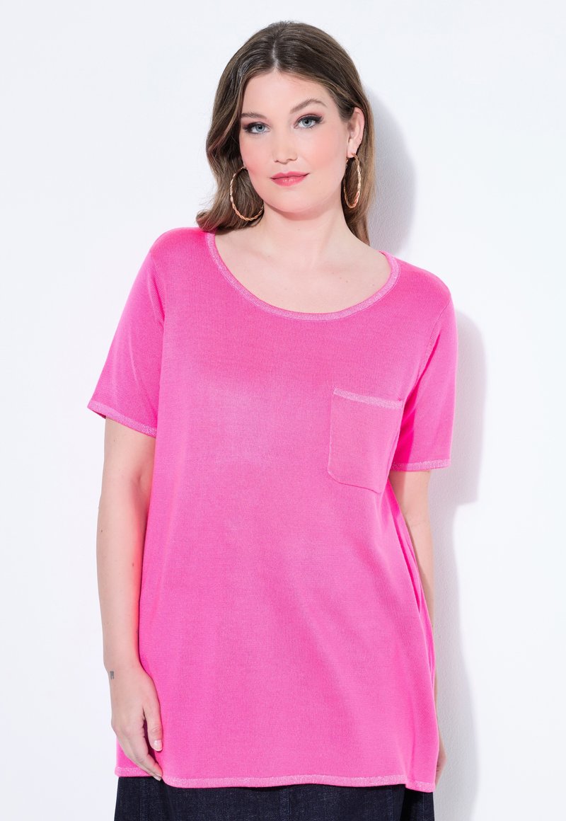 Frau mit langen braunen Haaren, die große Creolenohrringe trägt und ein hellpinkes T-Shirt mit kurzen Ärmeln sowie einer kleinen Brusttasche anhat.