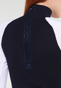 Haut tricot navy et blanc avec col monté ; présente une fermeture éclair navy brillante à l'arrière ; texture lisse avec des accents côtelés sur le col.