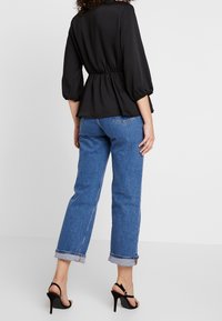 Black, flowy blouse with a cinched waist and three-quarter puff sleeves, paired with light blue cuffed straight jeans and black heeled sandals.