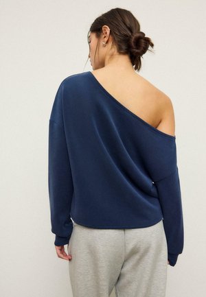 Ženska s temnimi lasmi v figi, oblečena v temno modro, dolgoročno bluzo brez ramen in svetlo sive hlače, obrnjena stran od gledalca na enobarvnem ozadju.