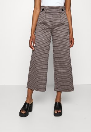 Vrouw draagt een broek met hoge taille, wijde pijpen en pied-de-poule motief met zwarte knoopdetails en zwarte plateau open teen sandalen, staand op een witte vloer.