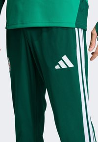 Zielone spodnie sportowe wykonane z gładkiej tkaniny, z białymi trzypasmowymi akcentami po bokach i białym logo Adidas.