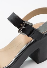Bride de cheville en cuir noir avec boucle argentée sur chaussure beige à talon haut avec semelle noire épaisse et talon bloc.