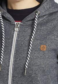 Sudadera con cremallera gris de tela texturizada, cordones en blanco y azul marino, cremallera plateada y un pequeño parche de logo marrón en la parte frontal.