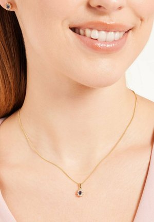 Collier en or avec un pendentif rond comportant une pierre précieuse bleue centrale entourée de petites pierres claires, porté sur la peau nue.